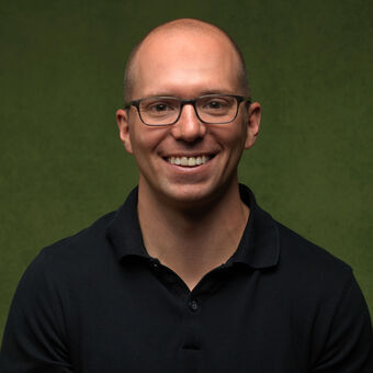 Portraiture of Adam DeLong. He is short, pale, and bald, but incredibly handsome. He is wearing glasses and facing forward against a green backdrop.